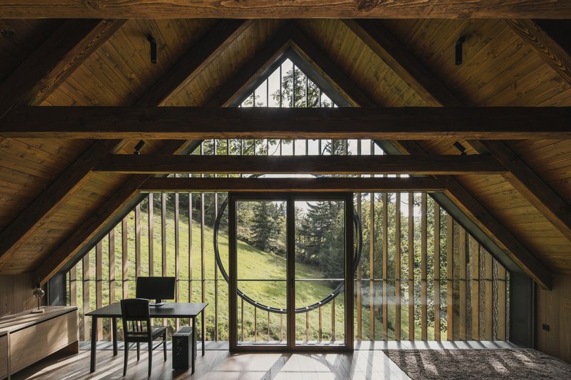 Interior loft space with exposed timber trusses and floor-to-ceiling glazing overlooking a hillside meadow