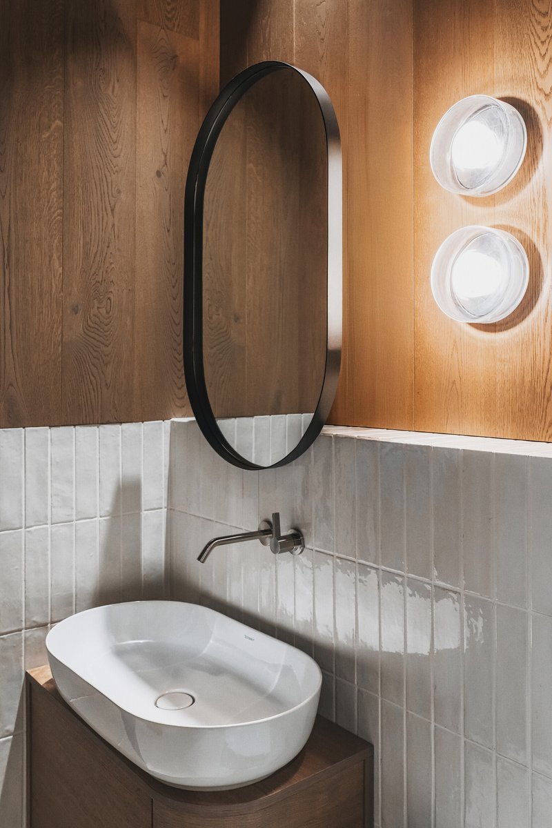 Oval vessel sink on timber vanity beneath vertical white tile and an oval mirror