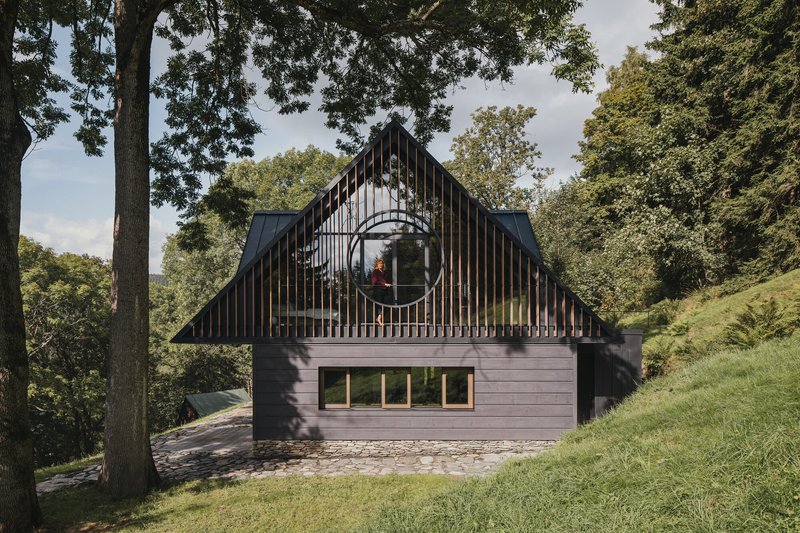 Gabled facade with vertical timber slat screen and circular window framed by mature deciduous trees