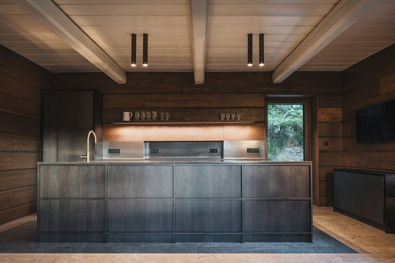 Kitchen island with dark steel cabinetry beneath exposed timber beams and recessed shelf lighting