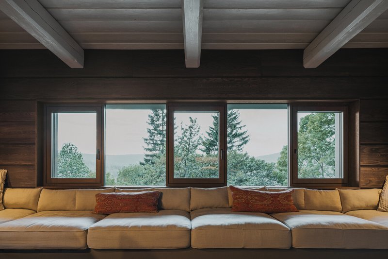 Five-panel window wall framing misty forest views above a seating area with exposed ceiling beams
