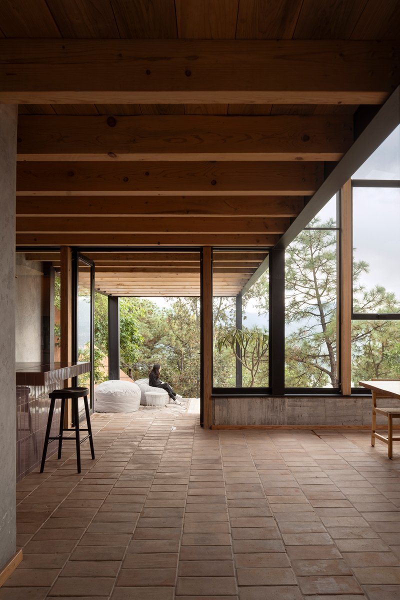 Clay tile floor terrace with exposed timber beams and steel-framed glazing looking toward forested hillside