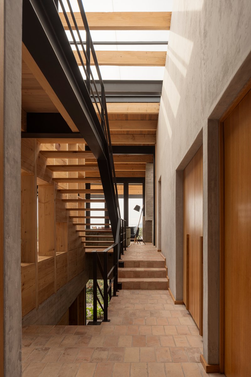 Corridor with open timber stair beside concrete walls and clay tile flooring under natural skylighting