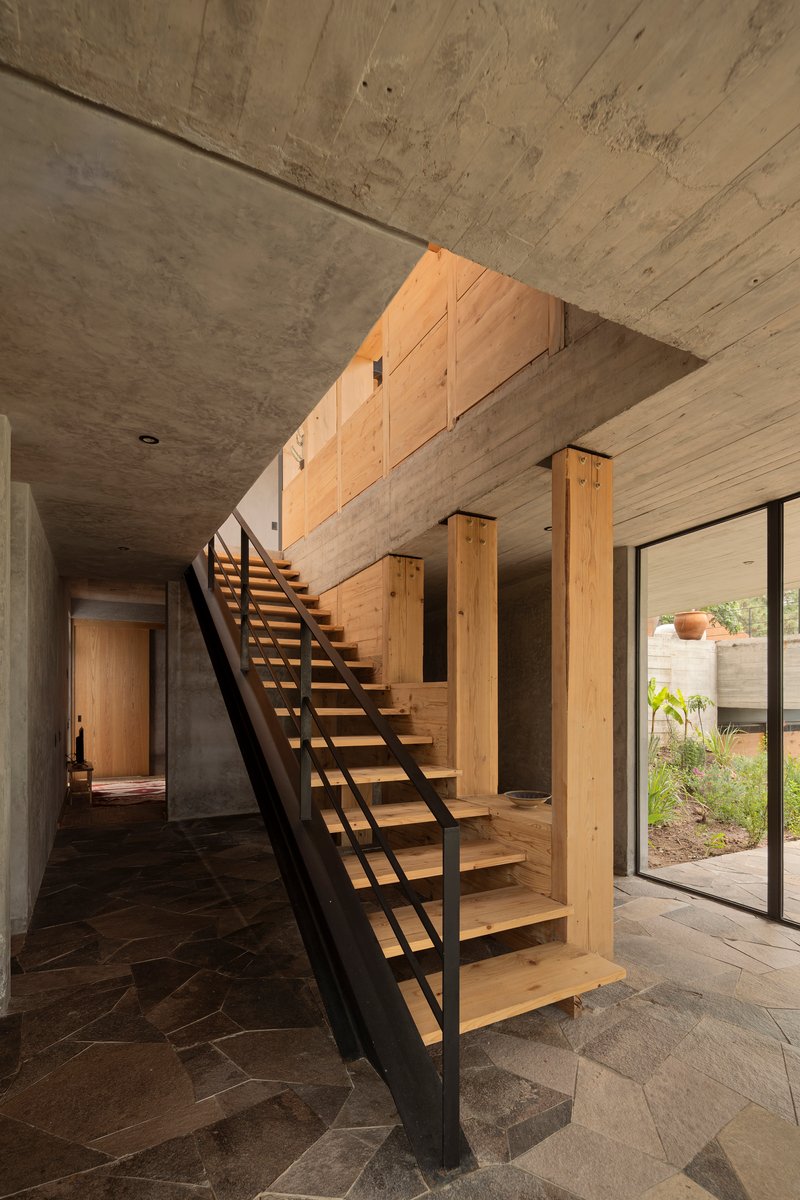 Open-tread timber staircase with black metal stringers in a double-height space with board-formed concrete walls