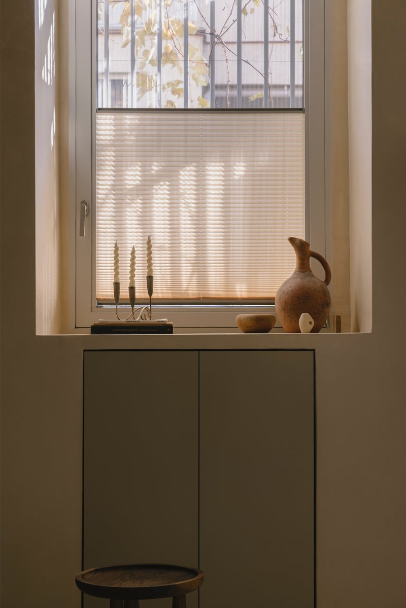 Window sill with pleated shade and terracotta jug catching afternoon sunlight above minimal cabinetry