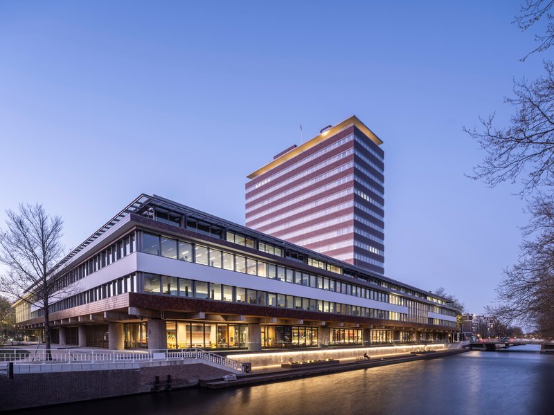 Canalside view of the horizontal building and illuminated tower at dusk with bare winter trees