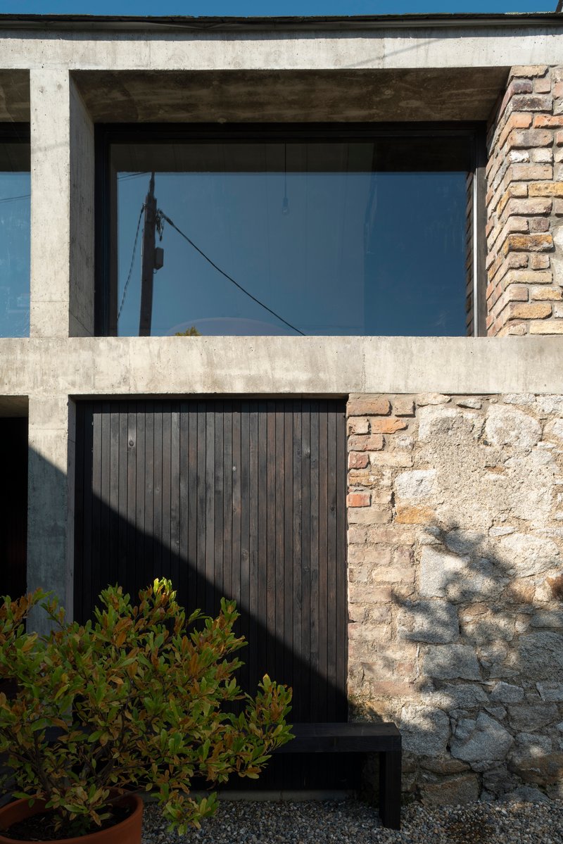 Layered facade showing concrete frame, black timber cladding, weathered stone wall, and planted shrubs in shadow