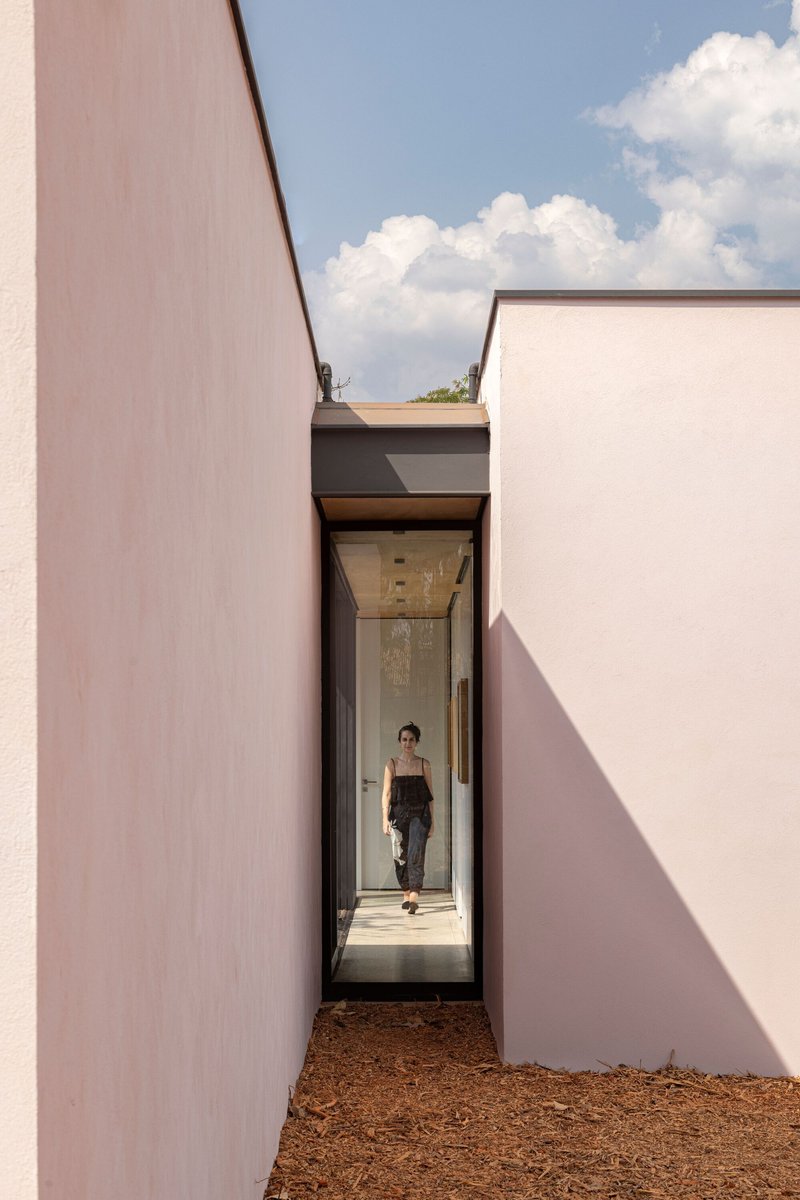 Narrow glass entry corridor framed by pale pink stucco walls with a figure walking through under blue skies