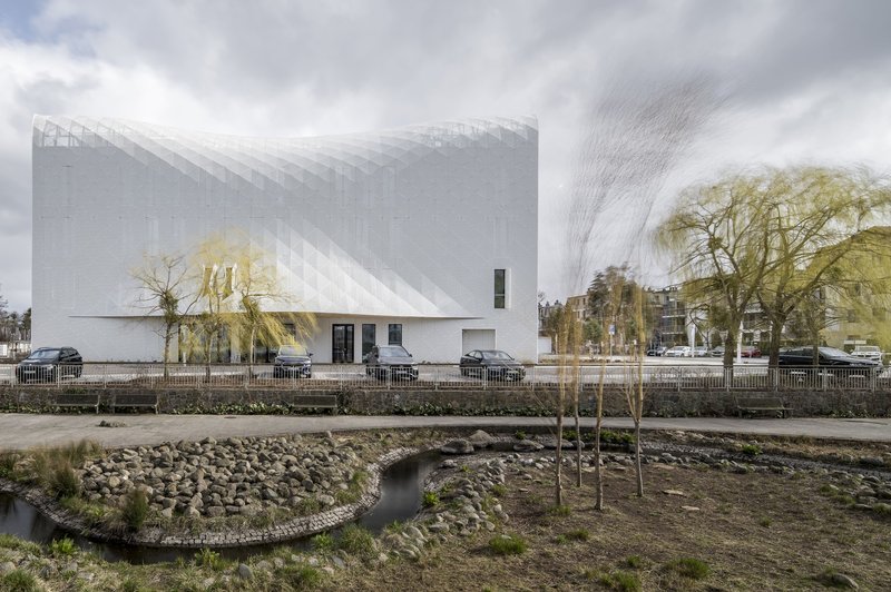 Street view of the textured white facade and curved volume across from a landscaped stream with young trees