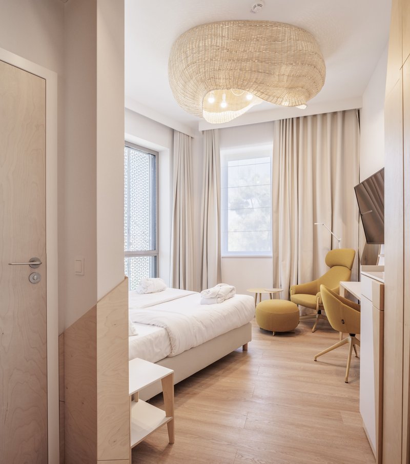 Bedroom with light wood flooring, woven pendant light, yellow armchair and curtained windows