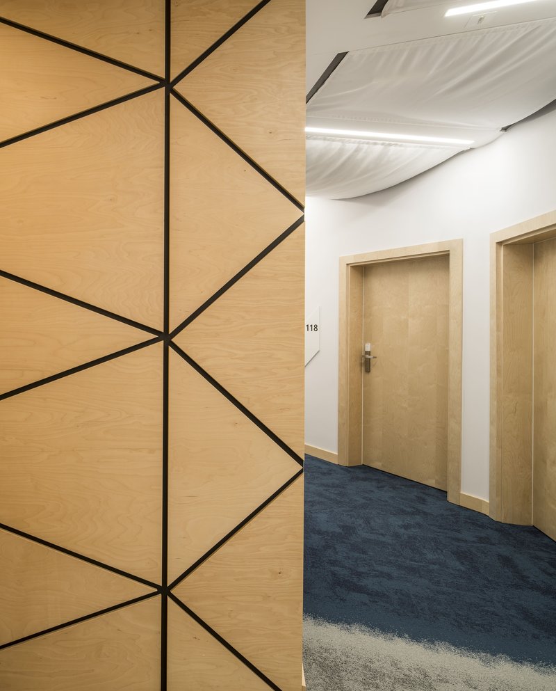 Corridor with geometric plywood wall panels and blue carpet leading to numbered guest room doors