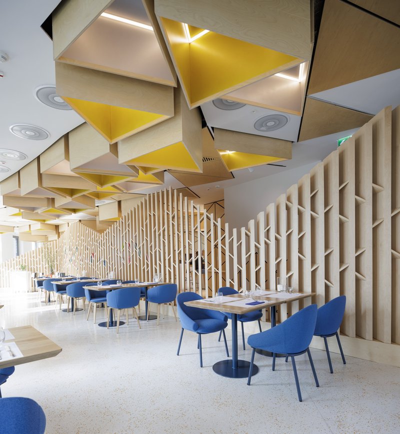 Interior dining area with folded plywood and yellow acoustic ceiling panels above blue upholstered chairs