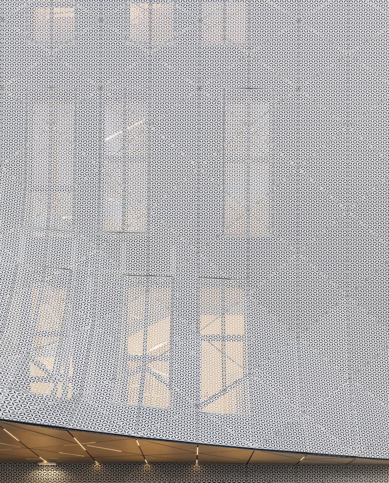Perforated metal screen wall with timber beams visible through the translucent surface at dusk