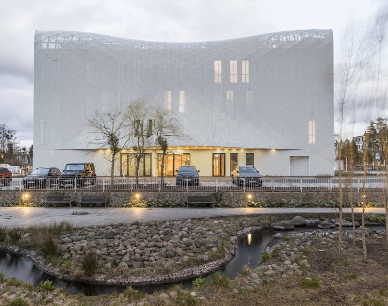 Curved perforated metal facade with illuminated base and reflecting pool at dusk
