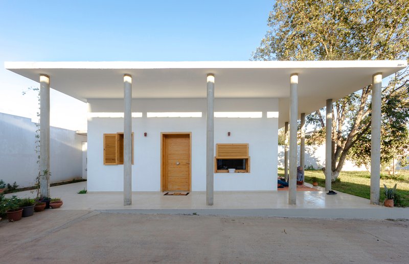 White stucco volume with wood shutters and central door beneath a flat canopy supported by concrete columns