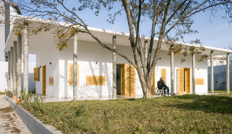 White facade with wood louvered windows and person in wheelchair on grass under late afternoon sun