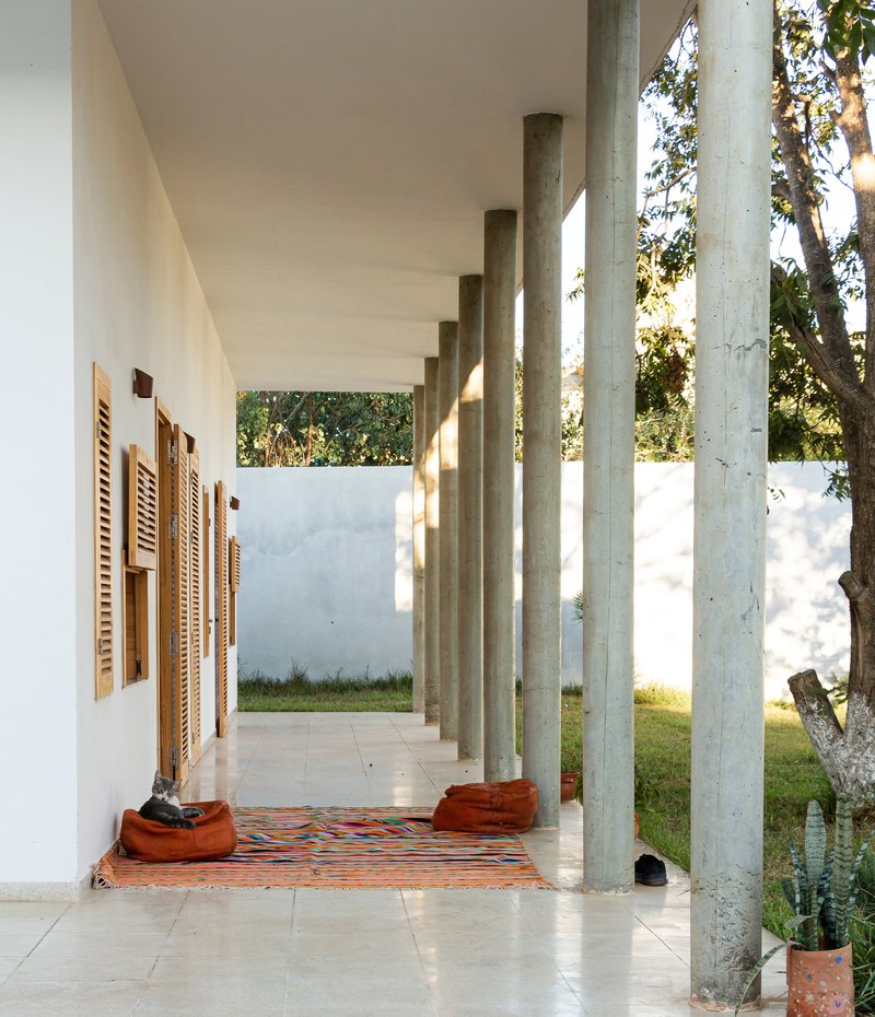 Covered walkway with concrete columns casting shadows over patterned textile rug on tile floor