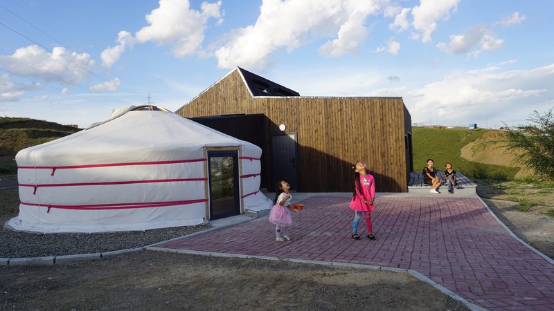 Courtyard terrace with brick paving where children play between the yurt and timber-clad volume at dusk