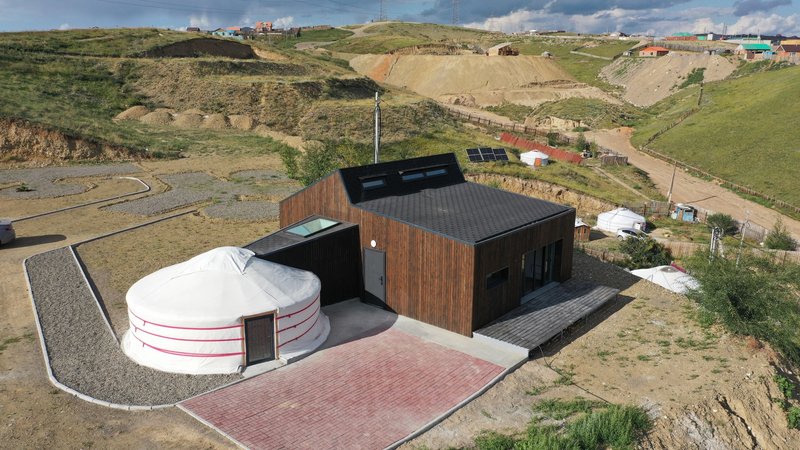 Aerial view of a circular yurt beside a timber-clad volume on a hillside site under scattered clouds