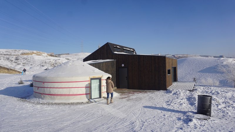 Dark timber-clad volume adjoining a circular yurt structure in a snow-covered landscape