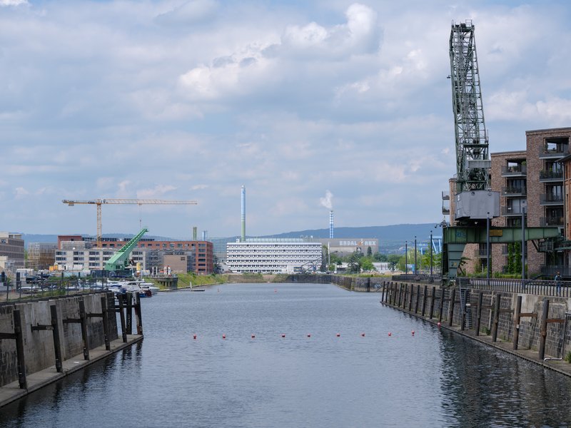 View across the canal showing the layered white facade with a crane and industrial chimney under scattered clouds