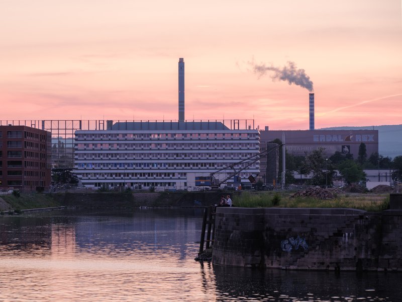 Horizontal facade reflected in the canal at dusk with a distant smokestack visible against the pink sky