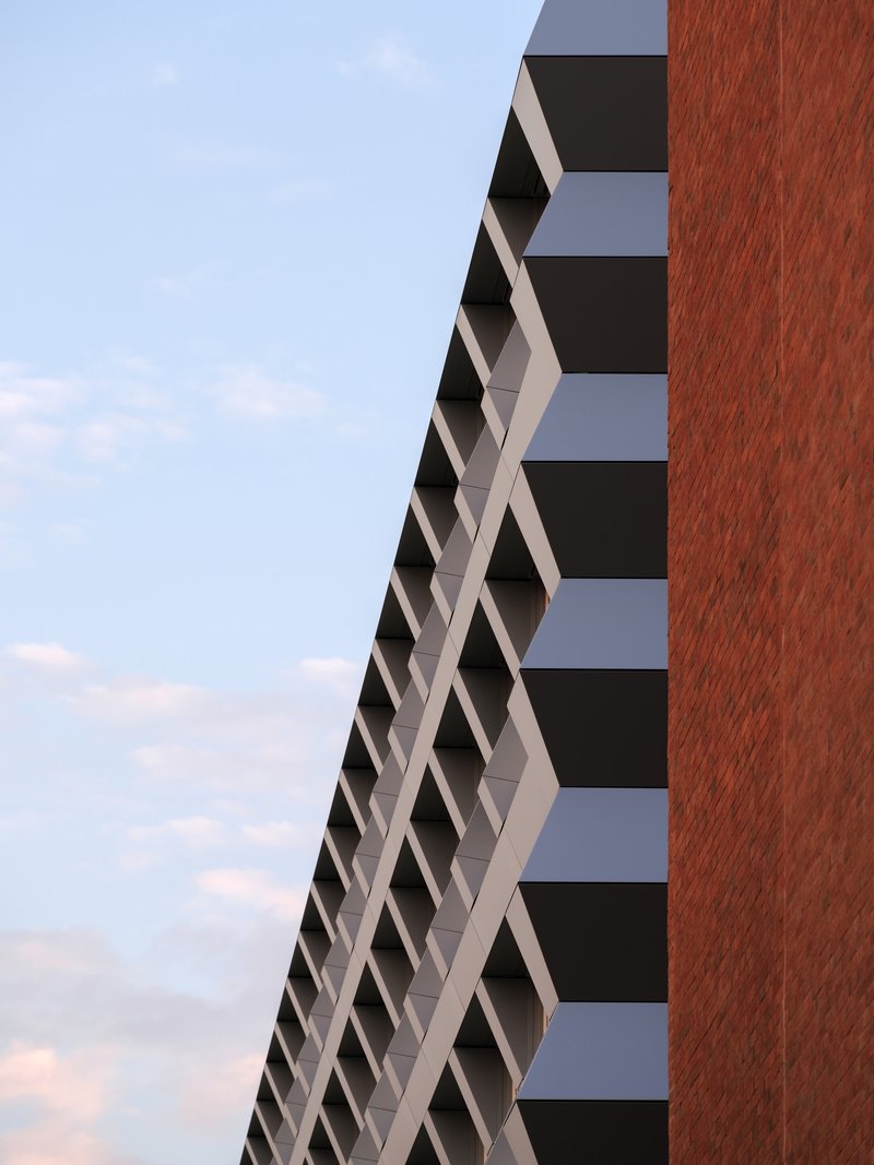 Diagonal facade with white sunshading elements meeting the vertical red brick tower volume at dusk