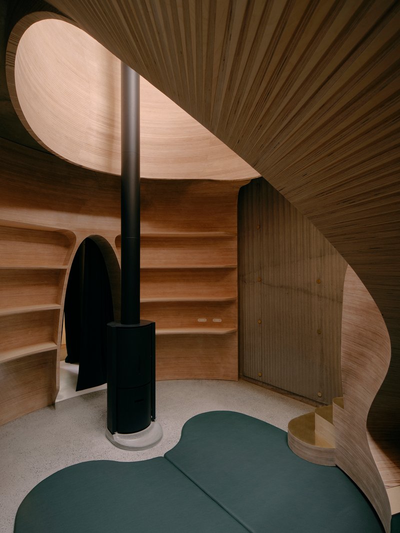 Curved timber interior space with black stove column and ribbed ceiling meeting organic timber walls