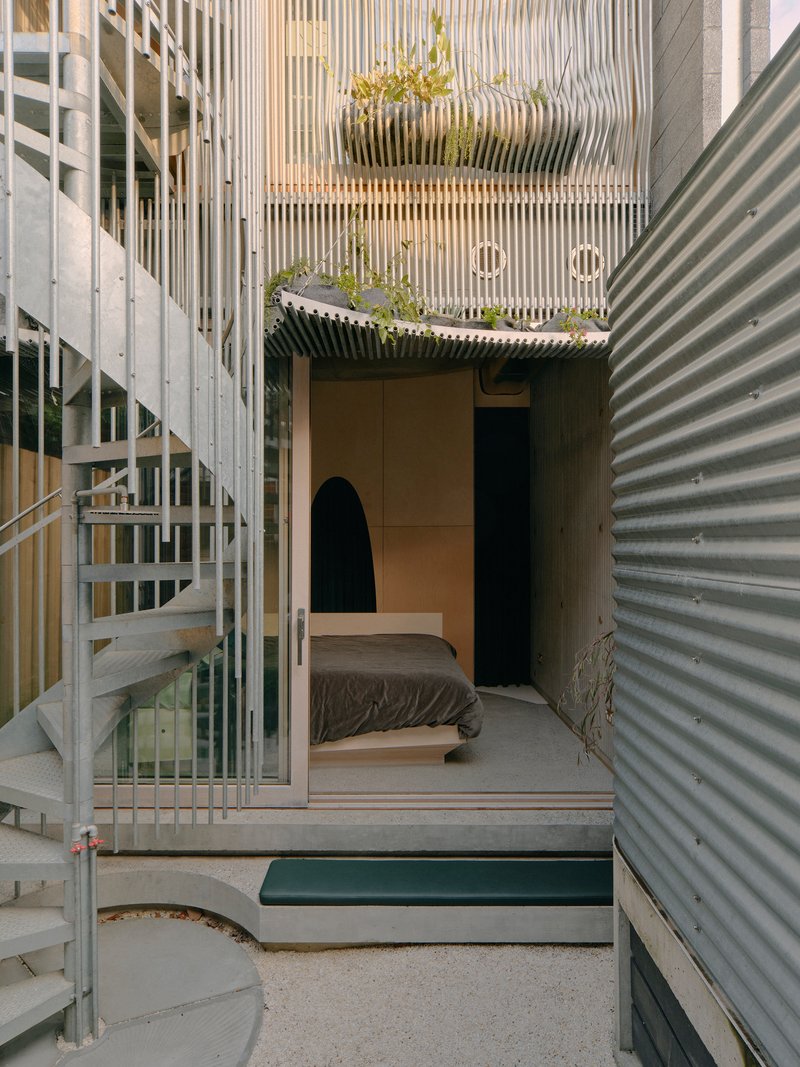 Courtyard view through vertical timber screen showing planted bed above corrugated metal walls and spiral stair