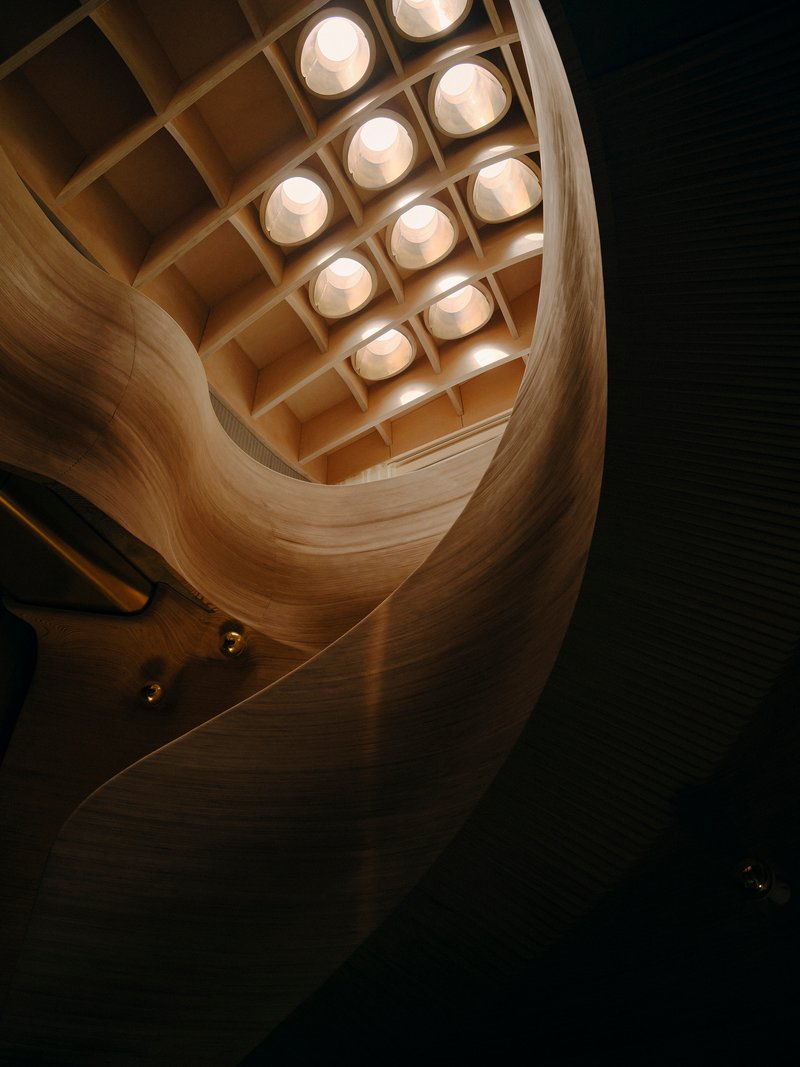 Upward view through curved timber stairwell with circular skylight coffered grid at dusk
