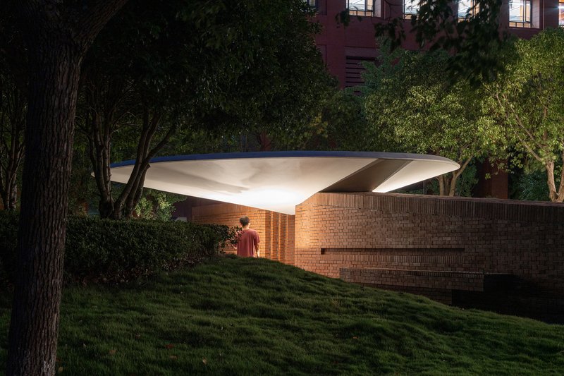 Illuminated canopy sheltering a brick pavilion wall as a figure stands on the lawn at twilight