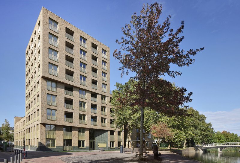 Tower volume in buff brick with deep balconies beside an autumn tree and canal edge