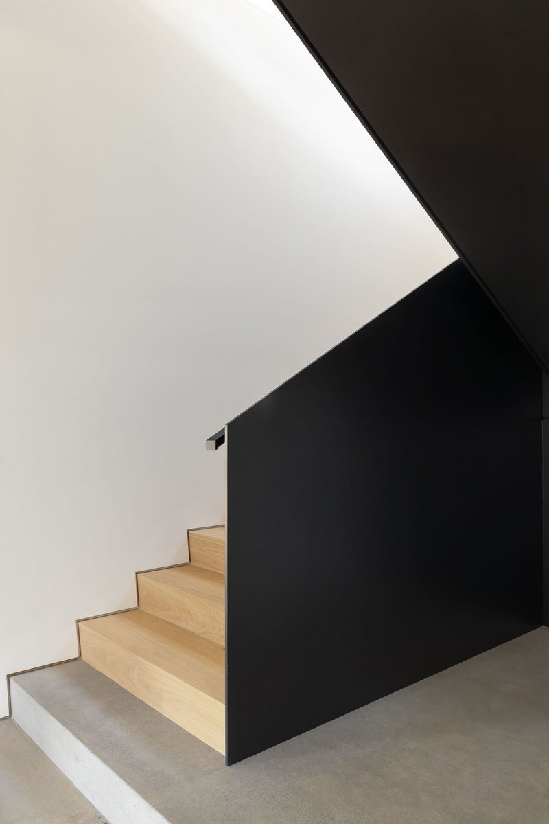 Staircase with light wood treads ascending alongside a black painted wall under a white vaulted ceiling