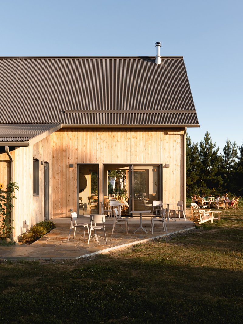 Light timber-clad facade with corrugated metal roof and glazed doors opening onto a terrace at sunset