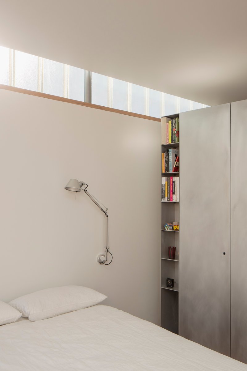 Bedroom with clerestory window above bed and narrow vertical bookshelf beside plywood cabinetry