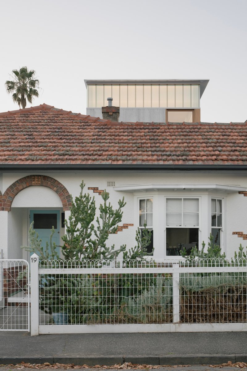 Street view of the white rendered bungalow with terracotta tile roof and metal-clad addition rising behind