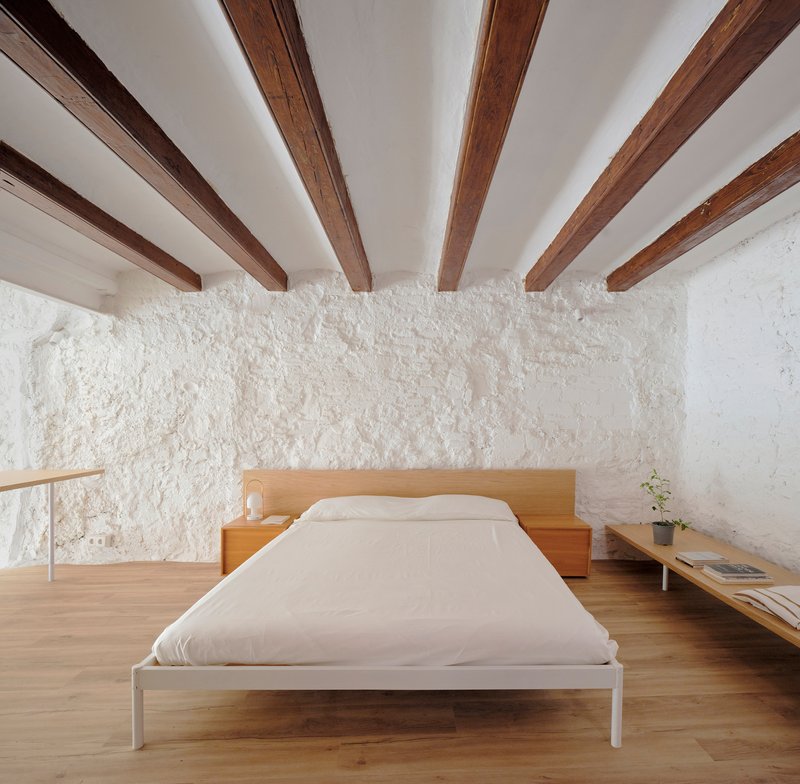 Bedroom with exposed timber ceiling joists above a timber platform bed against a rough white plaster wall