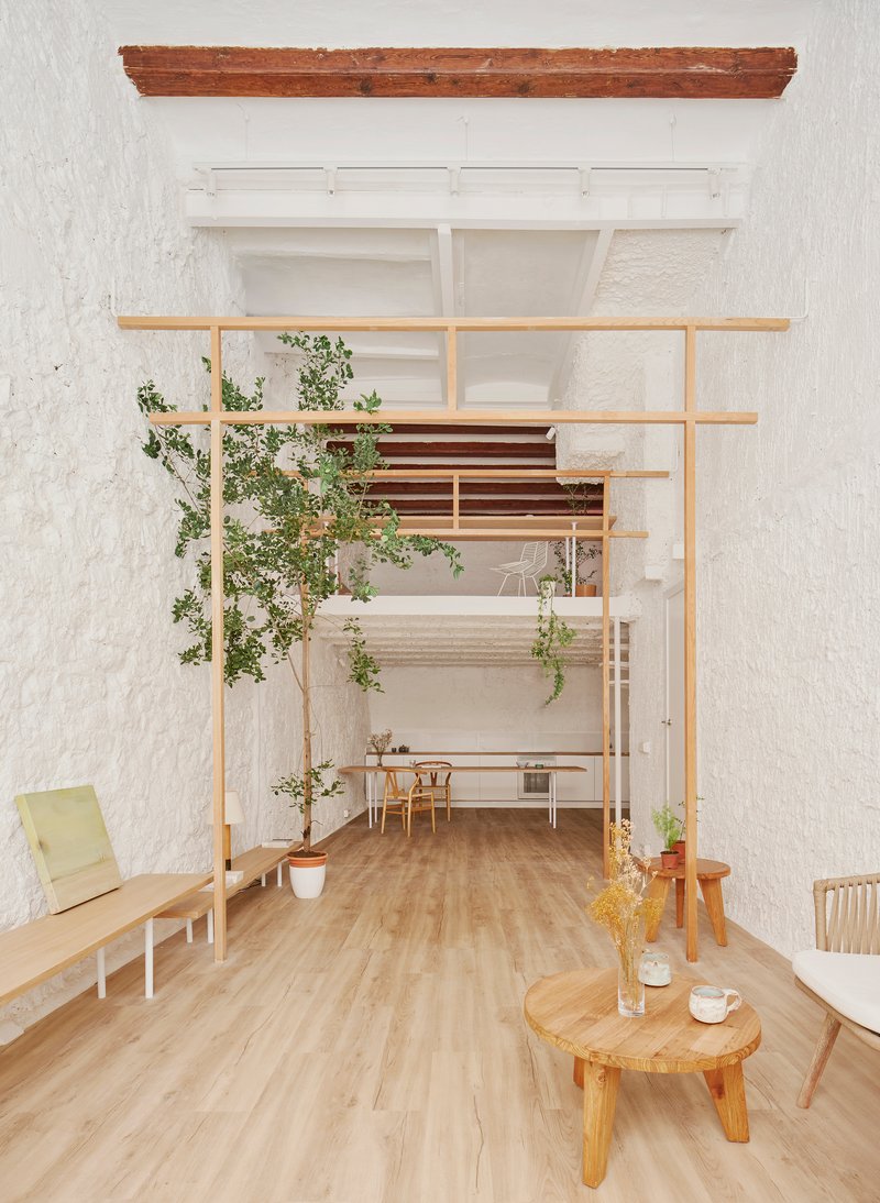 Double-height space with horizontal timber beams, exposed ceiling joists and a planted tree beside low furniture