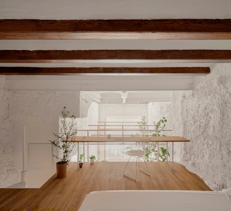 Double-height interior with exposed timber beams, timber mezzanine desk, and whitewashed textured walls with planted pots