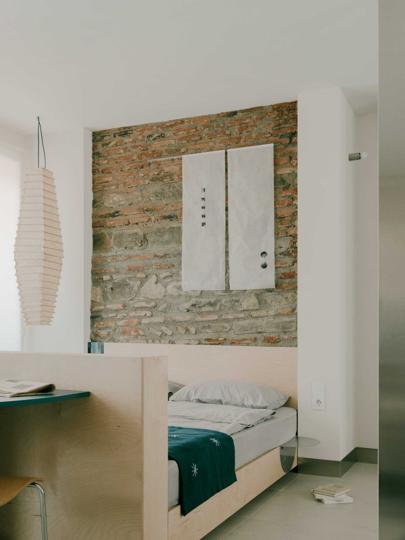 View across plywood platform bed to exposed brick feature wall with mounted white door panel