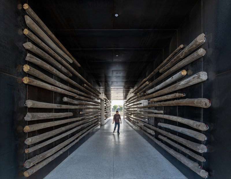 Corridor lined with reclaimed timber poles projecting from black walls as a visitor walks toward daylight