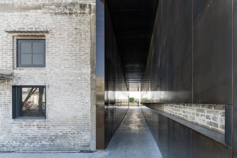 Passageway between weathered brick wall and blackened steel panel reflecting the opening ahead
