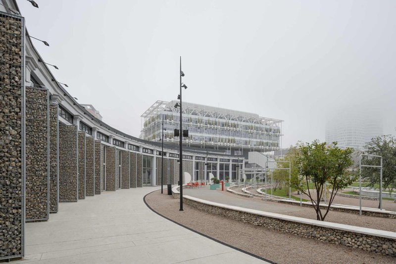 Curving path alongside a gabion wall leading toward the greenhouse under overcast sky