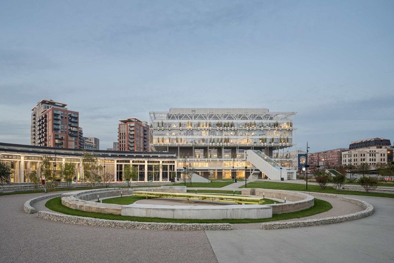 Circular sunken plaza with grass and gravel rings in front of the transparent greenhouse building at dusk