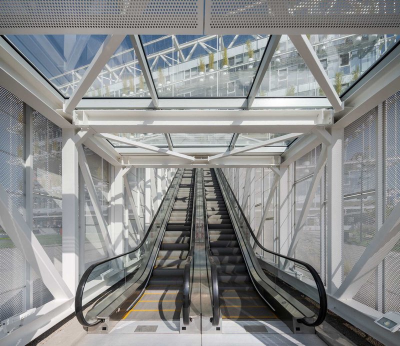 Escalator shaft enclosed by white steel trusses and perforated metal screens with views to courtyard beyond