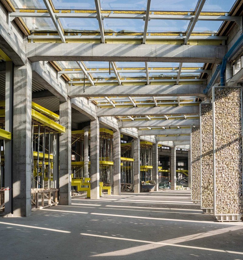Concrete columns and yellow steel beams beneath a glazed roof grid casting striped shadows