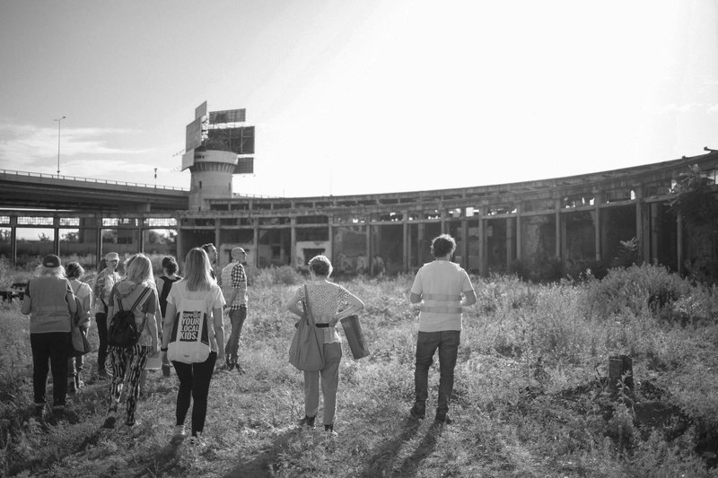 Group of visitors walking through overgrown grasses towards a curved colonnade and control tower at dusk