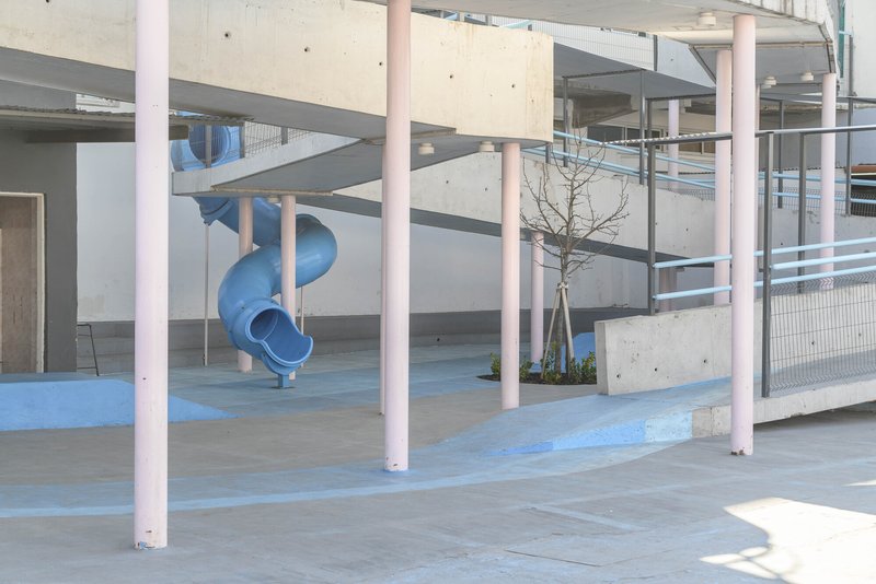 View through pink columns showing blue spiral slide and young planted tree with wire mesh railings