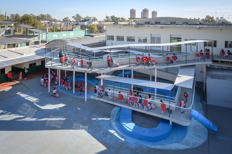 Elevated courtyard with blue tubular slide and schoolchildren in red uniforms gathering beneath corrugated metal canopies