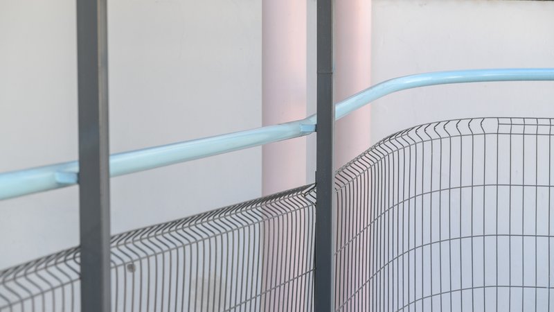 Close-up of curved wire mesh fence meeting blue metal handrail with pink columns in background